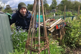 Man crouched down in an allotment. In the forefront of the shot is a wigwam for plants and veg to grow up.