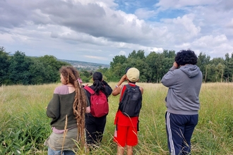 Group of youths out in nature using binoculars. They are standing in long grass in an open green space with trees and hills in the distance.