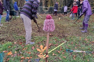 Image of a group at people at work in a woodland on the grassy floor. They are all doing garden work with spades and wheelbarrows.