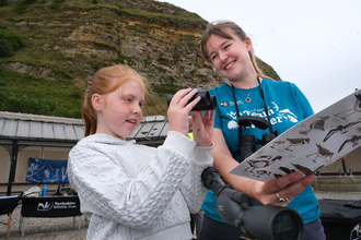 Child stood with binoculars looking out to see with a Yorkshire Wildlife Trust staff member showing her a marine wildlife spotting sheet.