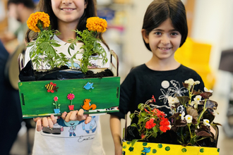 2 young girls smiling and holding their handmade planter boxes up to the camera. They have plants in and the wooden boxes are painted in yellow and green.