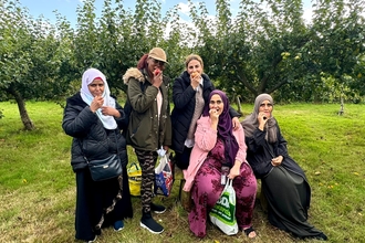 Group of ethnically diverse people stood and sat in a group eating appleas in an orchard.