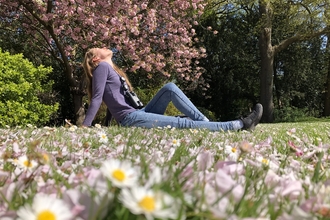 A woman sitting on the grass in a park, surrounded by flowers and trees, her face turned towards the sun