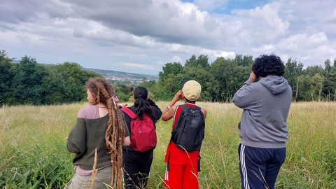 Group of youths out in nature using binoculars. They are standing in long grass in an open green space with trees and hills in the distance.