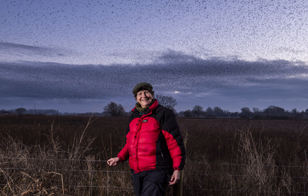 Woman standing on a nature reserve at dusk with a starling murmuraiton in the sky behind her. She is wearing a red winter coat and a dark coloured flat cap and smiling towards the camera. 