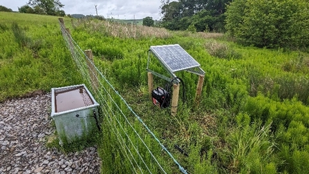 A solar panel (right) sits in a green field to the right of a fence, and a grey water trough (left)