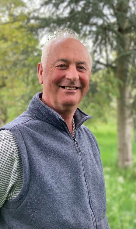 Professor Alastair Fitter in a blue bodywarmer taking a selfie in a woodland.