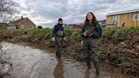 Two staff members in waders (centre) stand in a small brown stream with a riverbank behind.