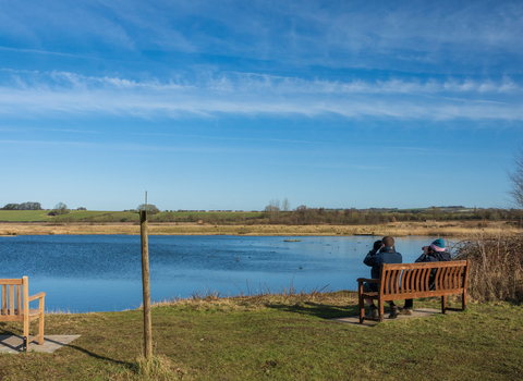 Two people sitting on a bench with binoculars looking out across the wetland lake on a nature reserve on a sunny cold day