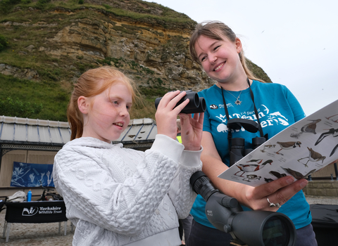 Child stood with binoculars looking out to see with a Yorkshire Wildlife Trust staff member showing her a marine wildlife spotting sheet.